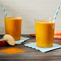 two glasses filled with orange juice next to carrots and an apple on a table