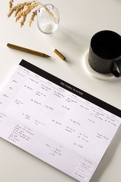 a coffee cup, pen and calendar sitting on a white table next to a vase