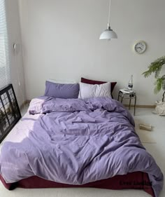 a bed with purple sheets and pillows in a white room next to a clock on the wall