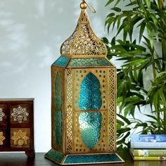 a green and gold lantern sitting on top of a wooden table next to a potted plant