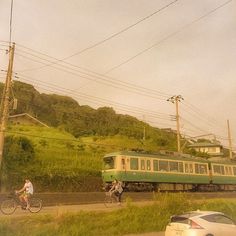 two bicyclists ride past a green train on the tracks in front of a car