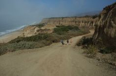 two people walking down a dirt road next to the ocean with cliffs in the background