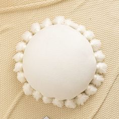 a round pillow with white pom - poms sits on top of a beige blanket