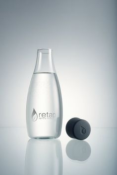 a glass bottle with a black cap next to it on a reflective surface, in front of a white background