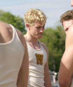 three young men standing next to each other in front of trees and grass, one wearing a white tank top