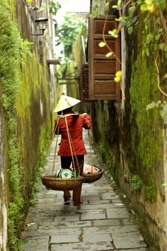 a woman walking down a narrow alley way carrying a basket full of food on her back