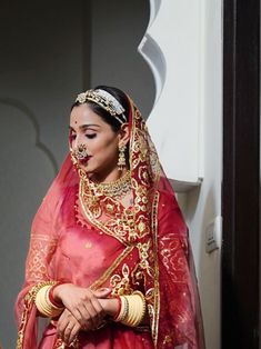 a woman wearing a red and gold bridal outfit with jewelry on her head, standing in front of a doorway