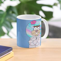 a blue coffee mug with a cartoon character on the front and bottom, sitting on a table next to a book