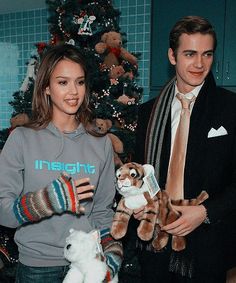 two people standing next to each other holding stuffed animals in front of a christmas tree