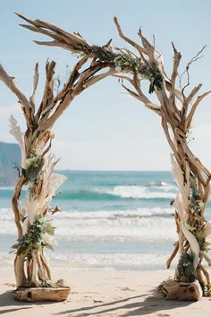 an arch made out of driftwood on the beach with flowers and greenery around it