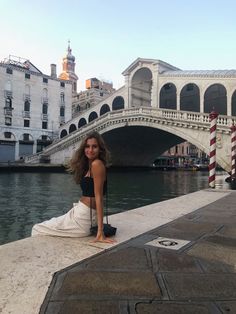 a woman sitting on the edge of a pier next to a river with buildings in the background