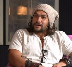 a man with long hair wearing a beanie and glasses sits in a chair looking at the camera