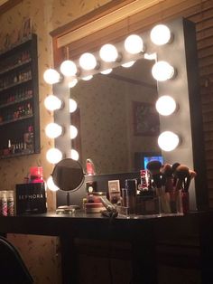 a vanity mirror with lights on it in front of a desk and shelves filled with cosmetics