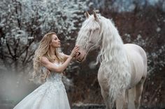 a woman in a wedding dress petting a white horse on the nose while it is snowing