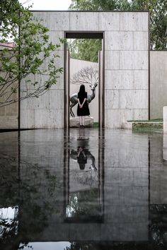 a woman standing in an open doorway with her reflection on the ground and trees behind her