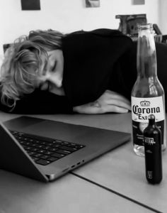 a woman laying her head on the table next to a beer bottle and laptop computer