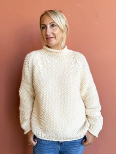 a woman standing in front of a pink wall wearing a white sweater and blue jeans