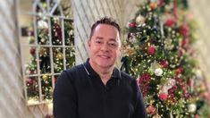 a man standing in front of a christmas tree with ornaments on it and smiling at the camera