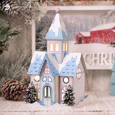 a small blue and white house with snow on the ground next to christmas tree branches