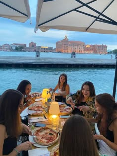 a group of women sitting around a table with pizzas on it in front of the water