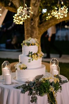 a white wedding cake sitting on top of a table next to a lit up tree