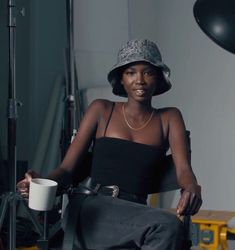 a woman sitting in a chair holding a coffee cup and wearing a camo hat