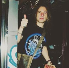 a man with long hair holding a pizza and giving the thumbs up