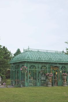 a green glass house in the middle of a grassy area with flowers and potted plants