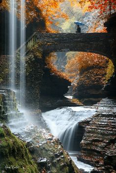 a waterfall in the middle of a forest with a bridge over it and a person standing on top