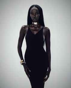 a woman in a black dress with gold jewelry on her neck and hands behind her back