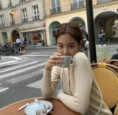 a woman sitting at an outdoor table drinking from a coffee cup in front of her