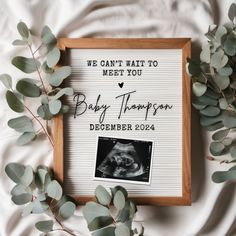 a baby announcement is displayed on a white sheet with eucalyptus leaves and a framed photo