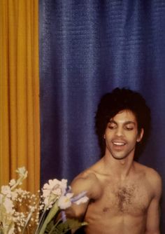 a shirtless man standing in front of a vase filled with flowers and looking at the camera