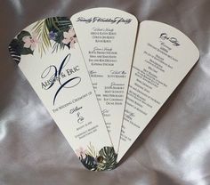 three fan shaped wedding programs on a silver satin surface with palm leaves and flowers in the center