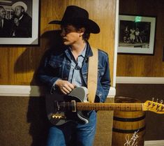 a man wearing a cowboy hat and holding a guitar in front of a wall with pictures on it