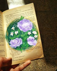 a hand holding an open book with flowers painted on the pages and in front of it