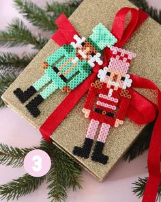 a cross stitch ornament hanging on a christmas present box with red ribbon and pine branches