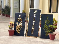 three black and gold welcome signs sitting on the ground next to potted plants in front of a building