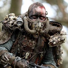 a man in a gas mask and full body armor walking through the woods with chains around his neck