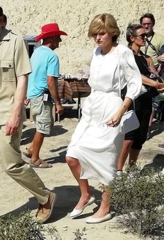 a woman in a white dress is walking on the beach with people watching from behind her