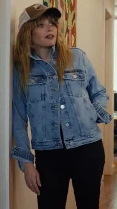 a woman wearing a denim jacket and cap standing in a hallway with her hands on her hips