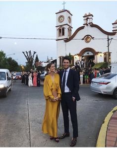 a man and woman standing in front of a church with cars parked on the street