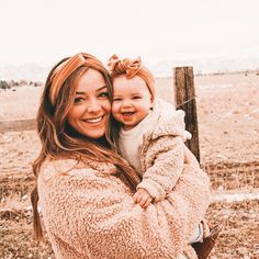 a woman holding a baby in her arms and smiling at the camera while standing next to a fence