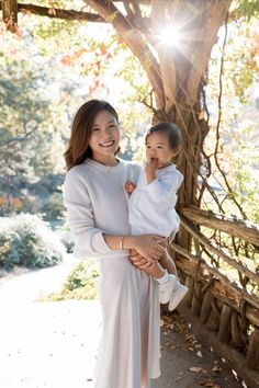 a woman holding a child in her arms while standing next to a tree with leaves on it