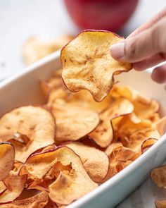 a person is dipping some potato chips into a white casserole dish with an apple in the background