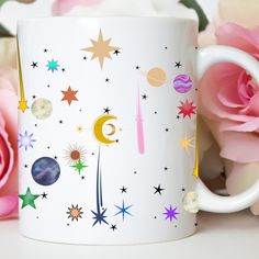 a white coffee mug with stars, planets and the moon on it next to pink roses