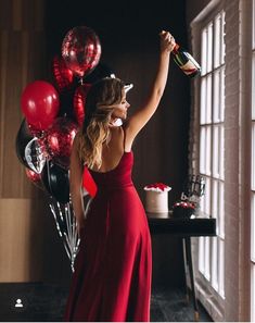 a woman in a red dress is holding a bottle and some black and red balloons