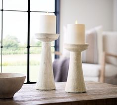 two white candles sitting on top of a wooden table next to a bowl and window