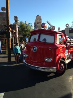 a red fire truck driving down a street with people standing around it and cars parked on the side of the road