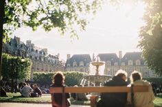 two people sitting on a bench in front of a building with a fountain behind them